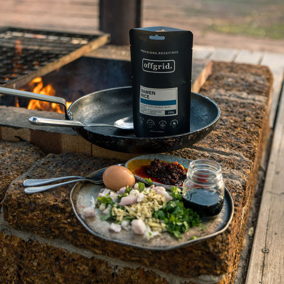 Outdoor cooking setup with a pan, jar, and food items on a rustic surface.